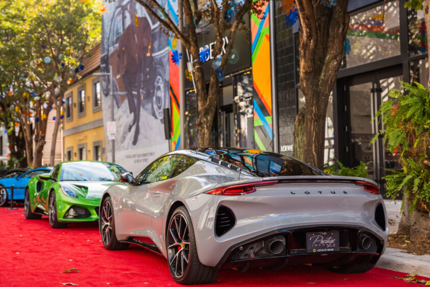 A silver Lotus sports car and a green car, both truly Driven By Design, are parked on a red carpet along a tree-lined street adorned with colorful murals and vibrant shops in the background. Lifestyle