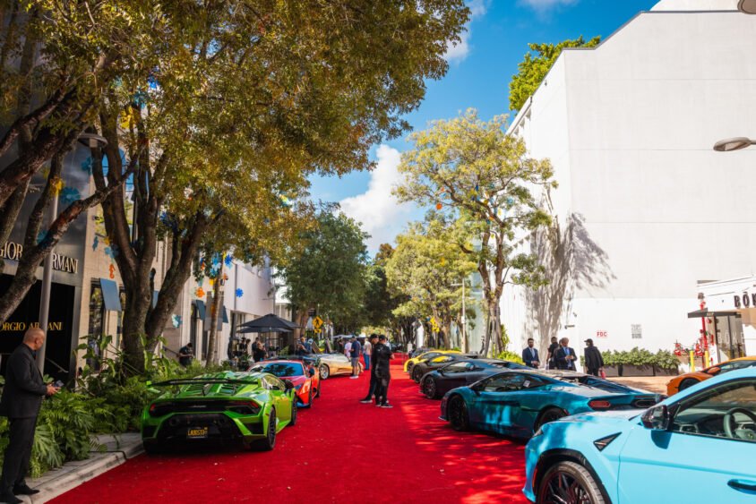 A row of colorful luxury sports cars, Driven By Design, lines a red carpet on a city street with trees and modern buildings under a bright blue sky. People admire the stunning cars and stroll nearby. Lifestyle