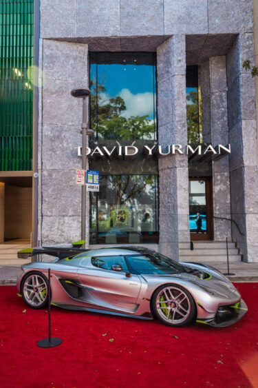 A silver sports car, Driven By Design, is parked on a red carpet in front of a modern stone building with large windows and a "David Yurman" sign above the entrance. Trees are reflected in the glass. Lifestyle