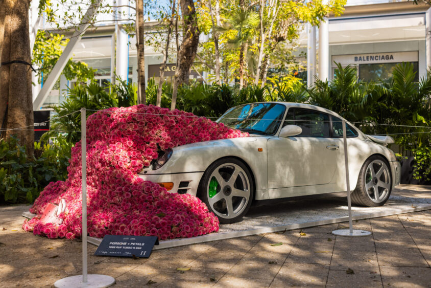 A white Porsche sports car is partly covered in a cascade of pink artificial roses, displayed outdoors near shops with a “Porsche + Petals” sign in front—an eye-catching scene truly Driven By Design. Lush greenery and storefronts fill the background. Lifestyle