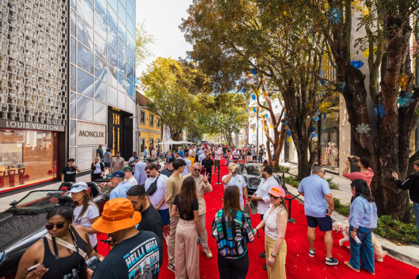 A bustling crowd walks along a red carpet on a tree-lined street with luxury stores like Louis Vuitton and Moncler, enjoying a lively outdoor event on a sunny day, all Driven By Design. Lifestyle