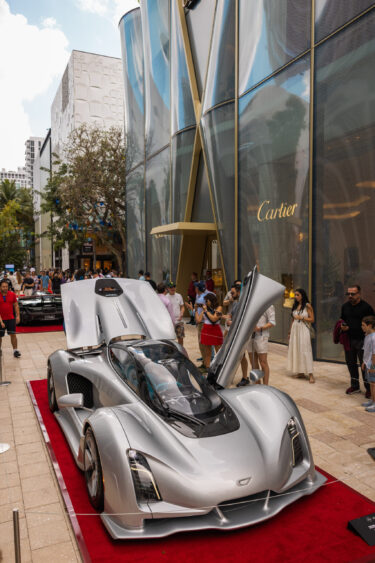A sleek silver sports car, Driven By Design, with its doors open is displayed on a red carpet outside a Cartier store, attracting a crowd of onlookers in a modern shopping district. Lifestyle