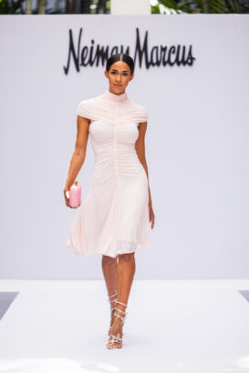 A woman walks a runway in a fitted, short-sleeve, light pink dress with ruching, lace-up heels, and a pink clutch, showcasing the latest fashion trends. The Neiman Marcus logo appears in the background. Lifestyle