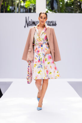 A woman models a floral dress with a pink blazer draped over her shoulders, embracing the latest fashion trends. She carries a matching floral bag and wears light blue heels, walking on a runway with a "Neiman Marcus" sign in the background. Lifestyle
