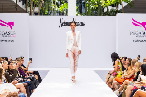 A model walks down a white runway at a fashion show, wearing a white blazer and light pink outfit. The audience sits on both sides, watching as Fashion takes the lead under the "Neiman Marcus" and "PEGASUS World Cup 2023" backdrop. Lifestyle