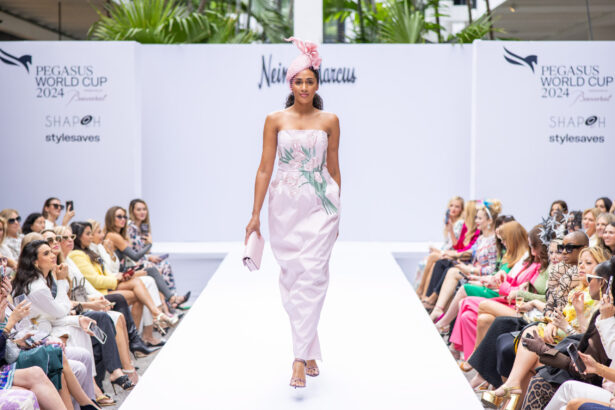 A model in a pale pink floral strapless jumpsuit and matching headwrap showcases the latest fashion trends on a white runway at a fashion show, with an audience on both sides and "Pegasus World Cup 2024" banners in the background. Lifestyle
