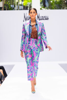 A model walks down a white runway showcasing bold fashion trends—a colorful suit with swirling pink, green, and turquoise patterns, a denim jacket, and a brown sequined top; the Neiman Marcus logo glows in the background. Lifestyle