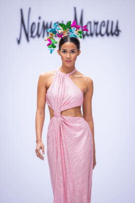 A model walks the runway showcasing fashion trends in a pink and white checkered cut-out dress and a colorful butterfly headpiece, with "Neiman Marcus" visible in the blurred background. Lifestyle