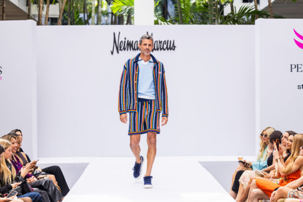 A male model walks a runway in a light blue shirt, navy sneakers, and a matching striped jacket and shorts, showcasing leading fashion trends. The audience sits on both sides as "Neiman Marcus" is displayed on the white backdrop behind him. Lifestyle