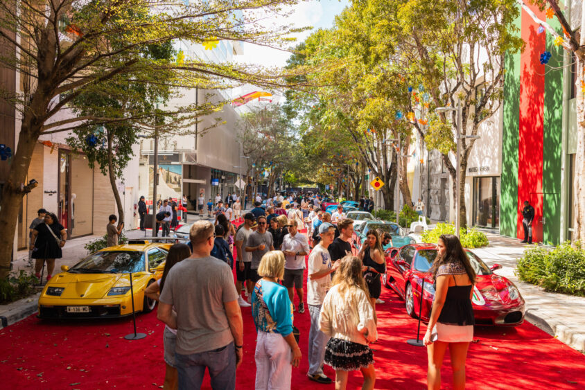 A crowd of people walks among luxury sports cars displayed on an outdoor red carpet along a tree-lined city street under a bright, sunny sky—an event truly Driven By Design. Lifestyle