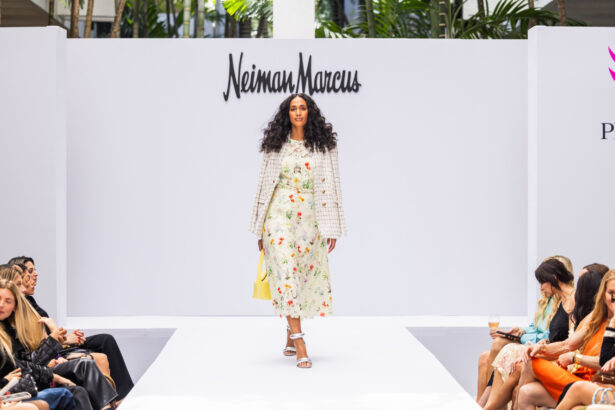 A model with long curly hair walks down a white runway wearing a floral dress, white jacket, and holding a yellow handbag, showcasing the latest fashion trends. The Neiman Marcus logo is displayed behind her as the audience watches. Lifestyle