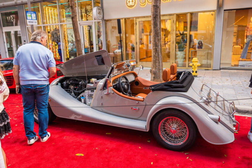 A classic silver convertible car, driven by design, sits with its hood open on a red carpet outdoors; a man inspects the engine while luxury shops line the background. Lifestyle