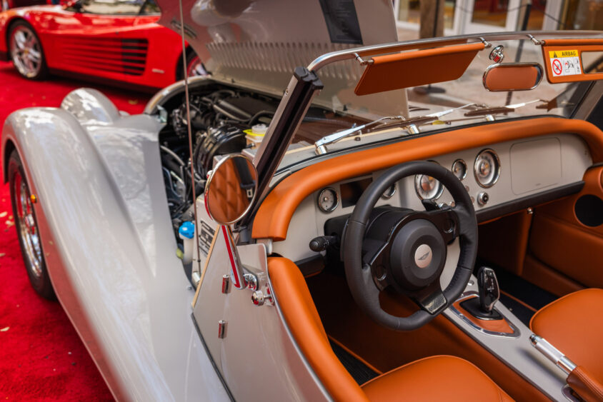 A close-up view of a vintage silver and orange convertible car, Driven By Design, with its hood open, showing the dashboard, steering wheel, and interior details on a red carpet. Another classic red car appears in the background. Lifestyle