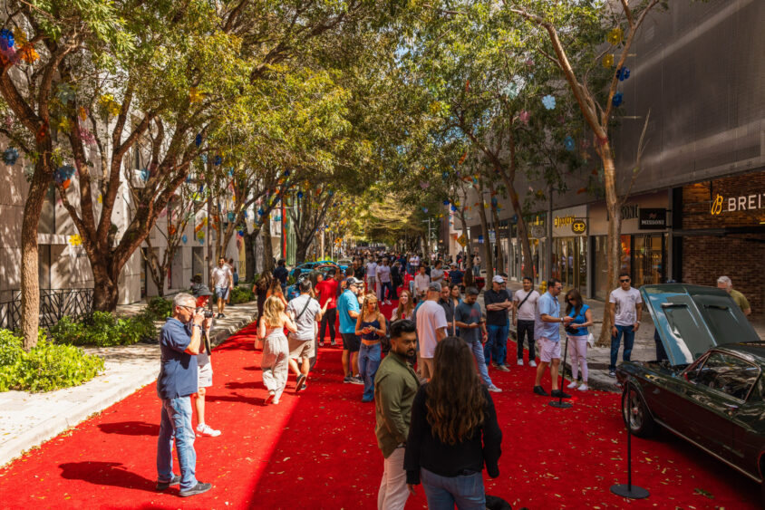 People gather on a red carpet-lined street filled with trees and luxury shops, viewing classic cars at the Driven By Design outdoor event and enjoying a sunny day surrounded by elegance. Lifestyle