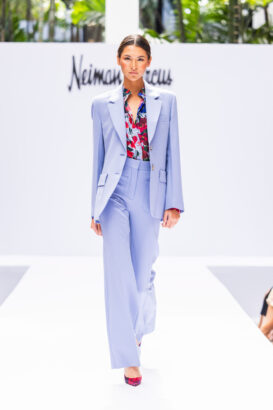 A model walks the runway wearing a light blue pantsuit with a colorful patterned blouse and red heels, proving that fashion takes the lead. The backdrop features the Neiman Marcus logo and greenery above a white runway setup. Lifestyle
