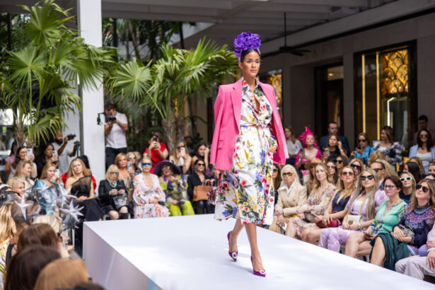 A model walks a runway outdoors, showcasing the latest fashion trends in a floral dress, bright pink blazer, purple heels, and a large purple headpiece. The audience, dressed stylishly, watches attentively from both sides of the catwalk. Lifestyle