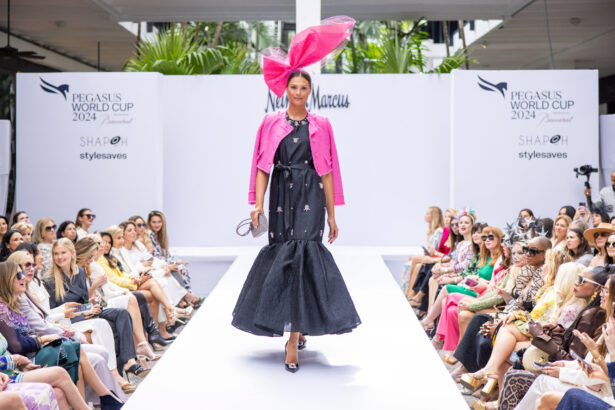 A model showcasing the latest fashion trends walks down the runway in a black dress, bright pink jacket, and large pink headpiece at an outdoor fashion show. The stylish audience snaps photos as "Pegasus World Cup 2024" signs are displayed. Lifestyle