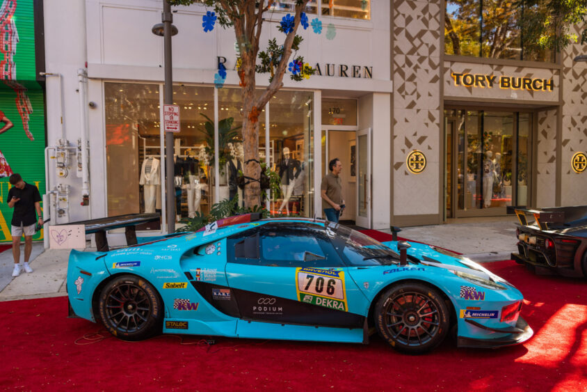 A turquoise race car with number 706, Driven By Design, is parked on a red carpet outside Ralph Lauren and Tory Burch stores, with people and trees visible in the background. Lifestyle