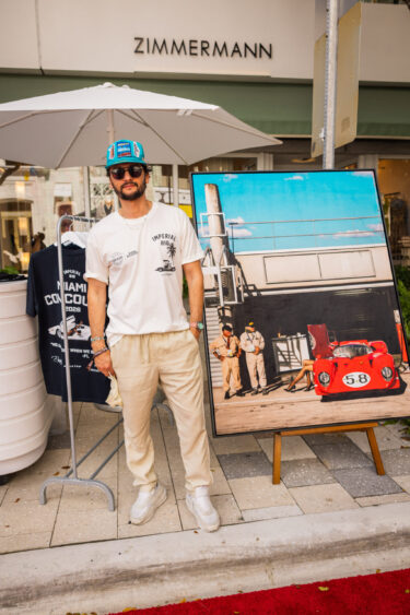 A man in sunglasses and a colorful hat stands beside a framed photo of a red race car and three people. Dressed casually, he poses with a white umbrella near the Zimmermann store, embodying the spirit of being Driven By Design. Lifestyle