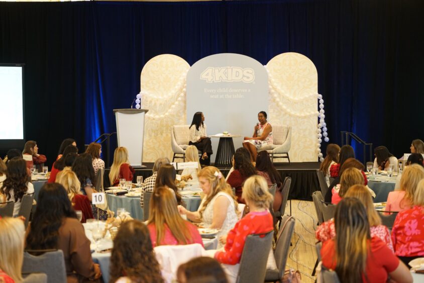 Two women sit on stage in conversation at a 4KIDS Broward Galentine’s fundraising event, while an audience of women at round tables watches attentively. A large 4KIDS logo and decorative backdrop are behind the speakers. Lifestyle