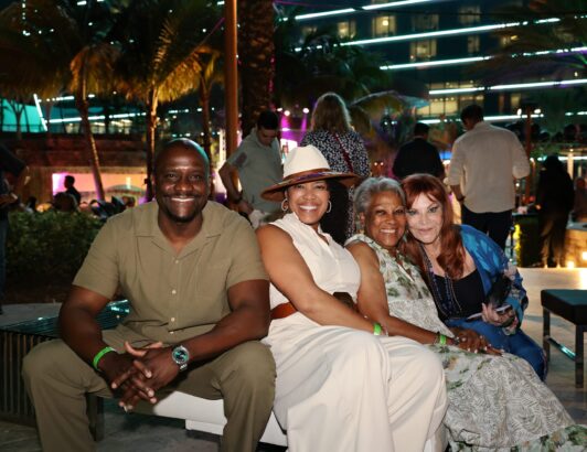 Four people sit closely together on an outdoor bench at night, smiling at the camera. Palm trees and colorful lights set a festive mood, capturing true Summer Vibes in this joyful moment. Lifestyle