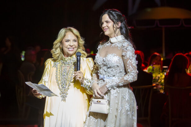 Two women stand together at a formal event. One holds a microphone and paper, wearing a light yellow gown with elaborate jewelry—a true Golden Angel. The other wears a sheer, floral-embroidered dress. Both are smiling, with people seated in the background. Lifestyle
