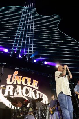 A band performs onstage at night in front of a large "Uncle Kracker" sign, while neon lights shaped like a giant guitar glow behind them, creating big impact and capturing summer vibes. Lifestyle
