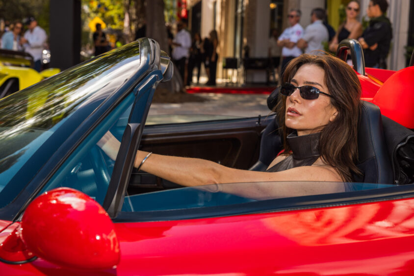 A woman wearing sunglasses sits confidently in the driver's seat of a red convertible, Driven By Design, with the top down and surrounded by people and luxury cars in the background on a sunny day. Lifestyle