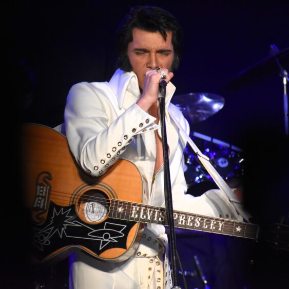 A performer dressed as Elvis Presley sings into a microphone while playing a guitar labeled "Elvis Presley," wearing a white jumpsuit with silver embellishments on a dimly lit stage during three shows over two nights. Lifestyle