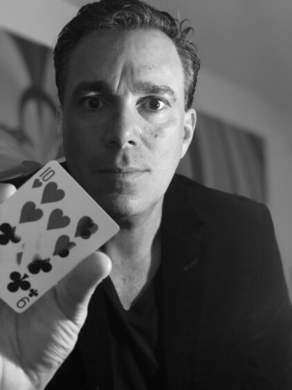 A man in a dark jacket holds up a playing card showing the 10 of hearts and 9 of clubs, looking seriously at the camera in a black and white photo—perfectly capturing the suspense before Three Shows, Two Nights. Lifestyle